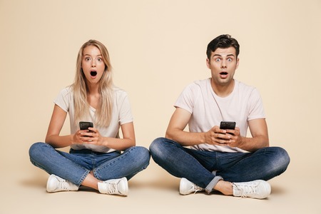 Portrait of a surprised young couple sitting together over beige background, using mobile phonesの写真素材