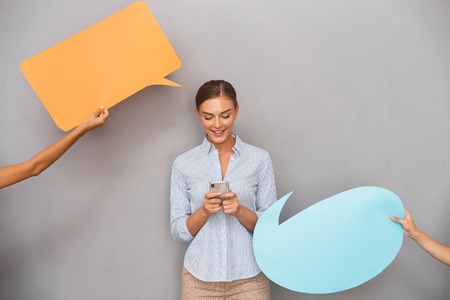 Image of beautiful business woman standing over grey wall background with speech bubbles chatting by...