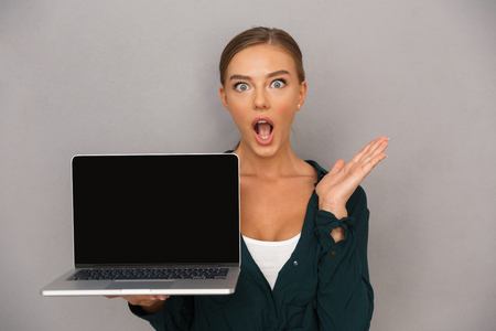 Shocked young businesswoman standing over gray background, showing blank screen laptop computerの写真素材