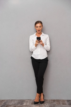 Image of beautiful shocked business woman standing over grey wall background using mobile phone.の写真素材