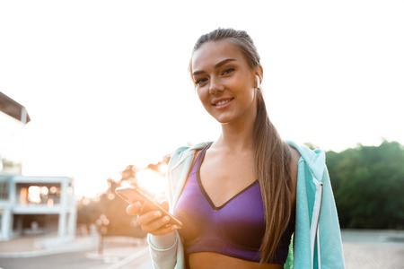 Photo of pretty cheerful young sports woman in park outdoors listening music with earphones using...