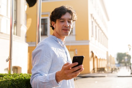 Handsome young casual man holding mobile phone while walking at the city street, listening to music...