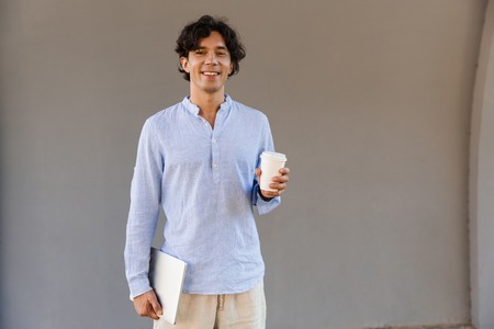 Smiling man dressed in shirt drinking coffee while standing outdoors at the city, holding laptop