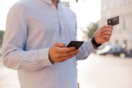 Cropped image of young casual man using mobile phone while walking at the city street, holding credit cardの写真素材