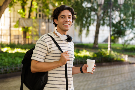 Smiling young casual man drinking coffee while walking at the city park, carrying backpackの写真素材