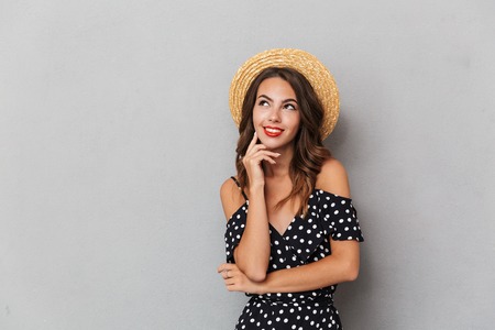 Portrait of a pensive young girl in dress and straw hat over gray background, looking away