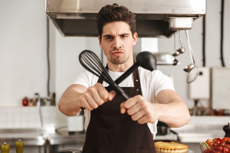 Furious young man chef cook in apron standing at the kitchen, holding ladle and whiskの写真素材