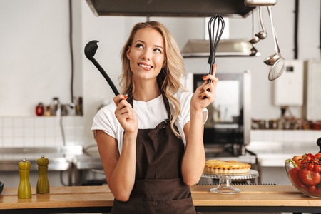 Smiling young woman chef cook in apron standing at the kitchen, holding ladle and whiskの写真素材
