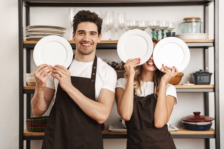 Image of happy excited young friends loving couple chefs on the kitchen holding plates covering faces.の写真素材