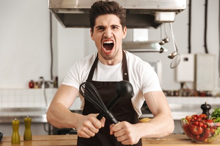 Angry young man chef cook in apron standing at the kitchen, holding ladle and whiskの写真素材