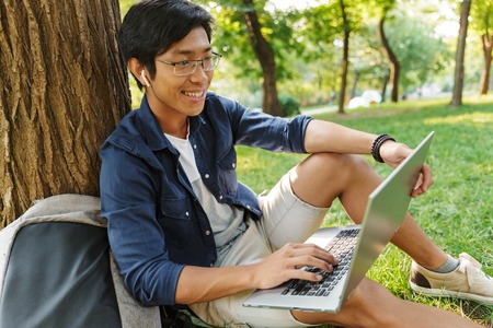 Side view of Smiling asian male student in eyeglasses using laptop computer while sitting near the tree in parkの写真素材