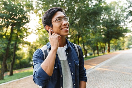 Smiling asian male student in eyeglasses and earphones listening music while looking away