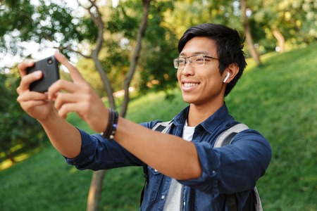 Smiling asian male student in eyeglasses making selfie on smartphone while being in parkの写真素材