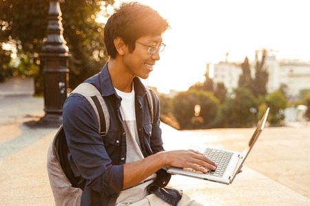Happy asian male student in eyeglasses using laptop computer while sitting outdoorsの写真素材