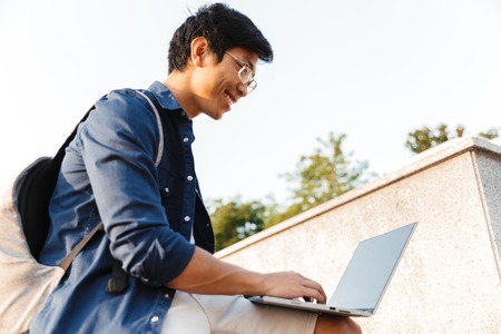 Low angle of smiling asian man student with backpack using laptop computer while sitting outdoorsの写真素材