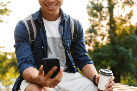 Cropped image of Smiling asian male student using smartphone and drinking coffee while sitting in parkの写真素材