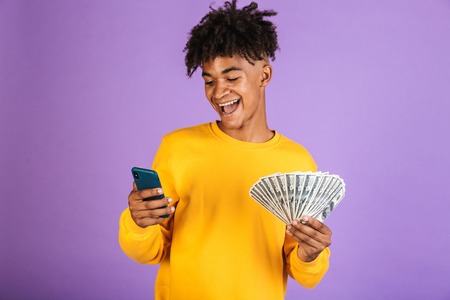Portrait of a happy young afro american man in sweatshirt isolated, showing money banknotes, using mobile phoneの写真素材