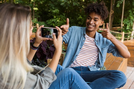 Excited young multiethnic couple spending time together at the park, taking photo of each other, showing thumbs upの写真素材