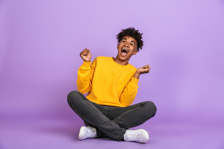Portrait of excited african american boy screaming and looking aside while sitting on floor with legs crossed isolated over violet backgroundの写真素材
