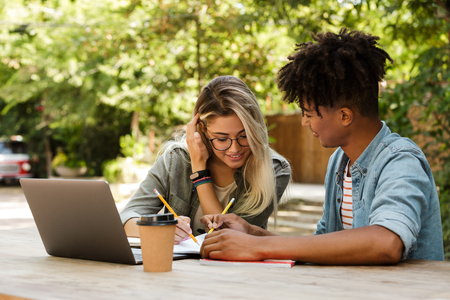 Lovely young multiethnic couple spending time together at the park, studying while sitting at the table with laptopの写真素材