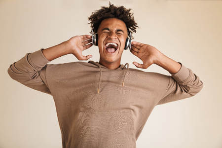Portrait of a happy young afro american man dressed in hoodie isolated, listening to music with headphones, screamingの写真素材