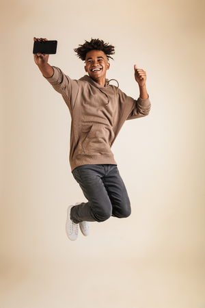 Full length portrait of a cheerful young afro american man dressed in hoodie taking a selfie isolated, showing thumbs upの写真素材
