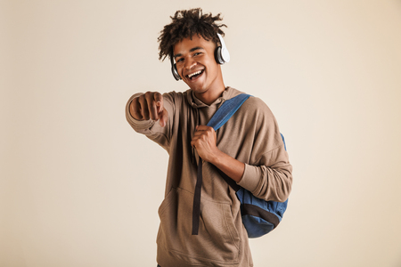Portrait of a cheerful young afro american man dressed in hoodie with headphones carrying backpack isolated, pointing at cameraの写真素材