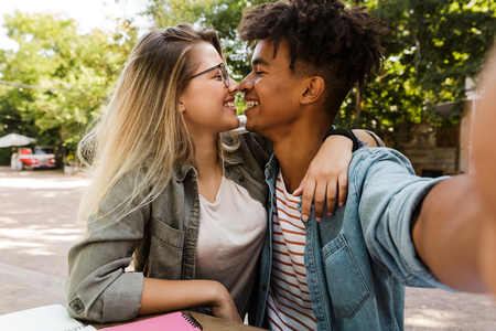Lovely young multiethnic couple spending time together at the park, studying while sitting at the table and taking a selfieの写真素材