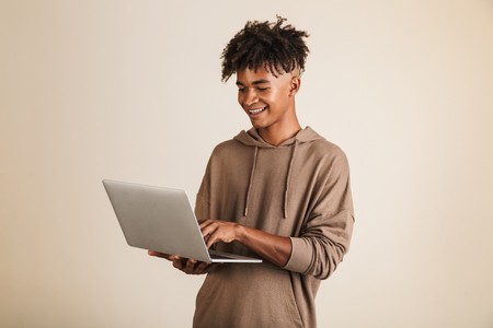 Portrait of a smiling young afro american man dressed in hoodie isolated, using laptop computerの写真素材