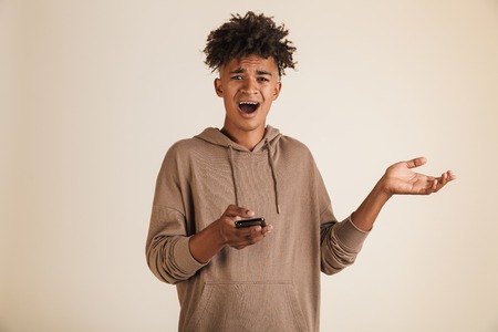 Image of a shocked confused young afro american man dressed in hoodie isolated, holding mobile phone looking camera.の写真素材