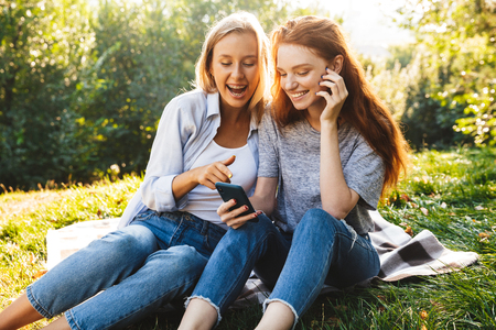 Image of pretty women students wearing casual clothes sitting on grass and using smartphone while resting in summer parkの写真素材