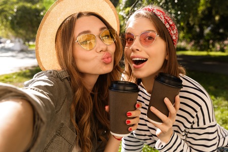 Two smiling young girls friends having fun at the park, taking a selfie, holding coffee cups