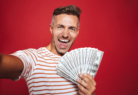 Photo of delighted man in striped t-shirt smiling and taking selfie while holding fan of money banknotes isolated over red backgroundの写真素材