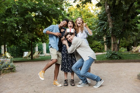 Image of happy young group of friends outdoors in park having fun holding gift box.