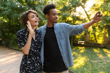 Image of a smiling young multiethnic loving couple standing posing in park outdoors hugging take a selfie by phone waving.の写真素材