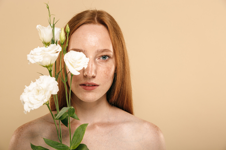 Beauty portrait of a lovely young topless redhead girl with freckles isolated over beige background, posing with eustoma flowersの写真素材