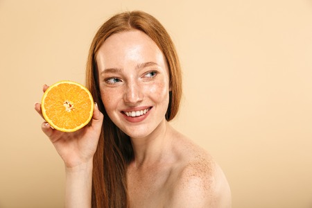 Beauty portrait of a smiling young topless redhead girl with freckles isolated over beige background, holding slice of an orangeの写真素材