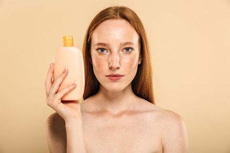 Image of pretty amazing young redhead woman standing isolated over beige background wall holding cream for skin care. Looking at camera.の写真素材
