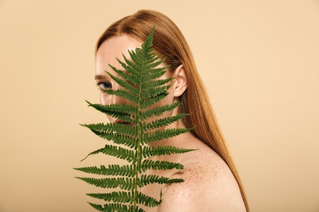 Image of beautiful pretty young redhead woman standing isolated over beige wall background holding leaf flower plant.の写真素材