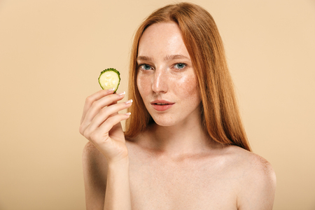 Beauty portrait of a beautiful young topless redhead girl with freckles isolated over beige background, holding slice of a cucumberの写真素材