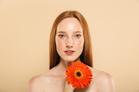 Photo of cute gorgeous young redhead woman standing over beige background wall looking camera holding flower.の写真素材