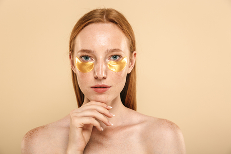 Beauty portrait of a beautiful young topless redhead girl with freckles isolated over beige background, wearing eye patch face maskの写真素材