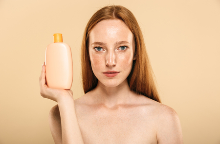 Image of pretty amazing young redhead woman standing isolated over beige background wall holding cream for skin care. Looking at camera.の写真素材