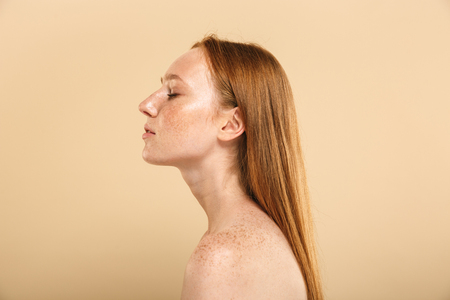 Beauty portrait of a young topless redhead girl with freckles isolated over beige background, side viewの写真素材