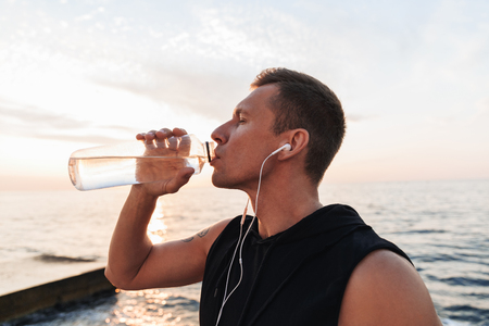 Picture of handsome strong young sportsman outdoors at the beach listening music with earphones drinking water.の写真素材