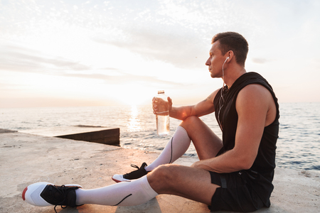 Picture of handsome strong young sportsman outdoors at the beach listening music with earphones drinking water.の写真素材