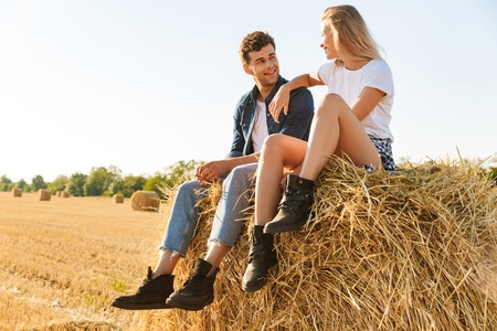 Smiling young couple sitting together on a haystack at the field, talkingの写真素材