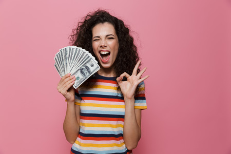 Image of joyous woman 20s with curly hair holding fan of dollar money isolated over pink background