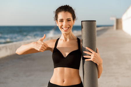 Confident young sportswoman standing outdoors at the seaside, holding fitness mat, showing thumbs upの写真素材