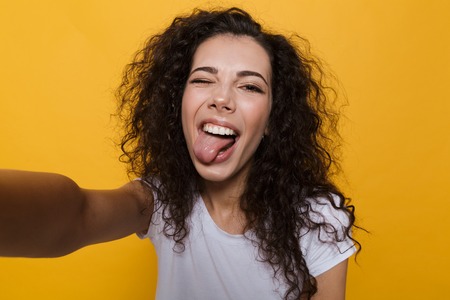 Image of an excited happy cute young woman posing isolated over yellow background take a selfie by camera showing tongue.の写真素材